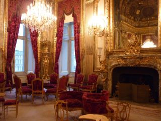 Salón del departamento de Napoleón III en el Louvre. Cortinas rojas, lámparas decoradas y un gran espejo arriba de la chimenea.